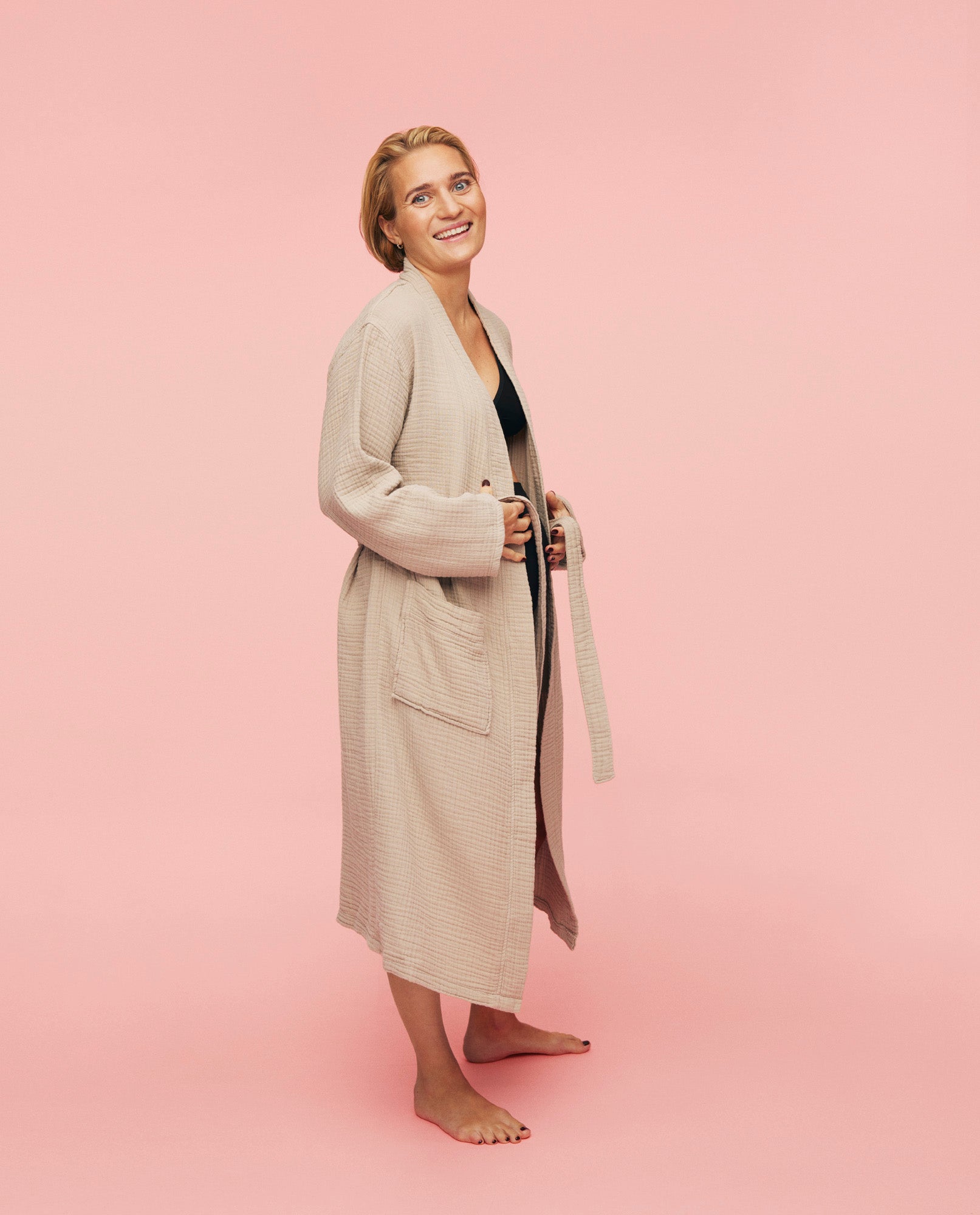 Kvinde smiler imens hun er iført momkind kind robe. Den er åben og hun holder fast i snoren som er til at lukke kåben med.