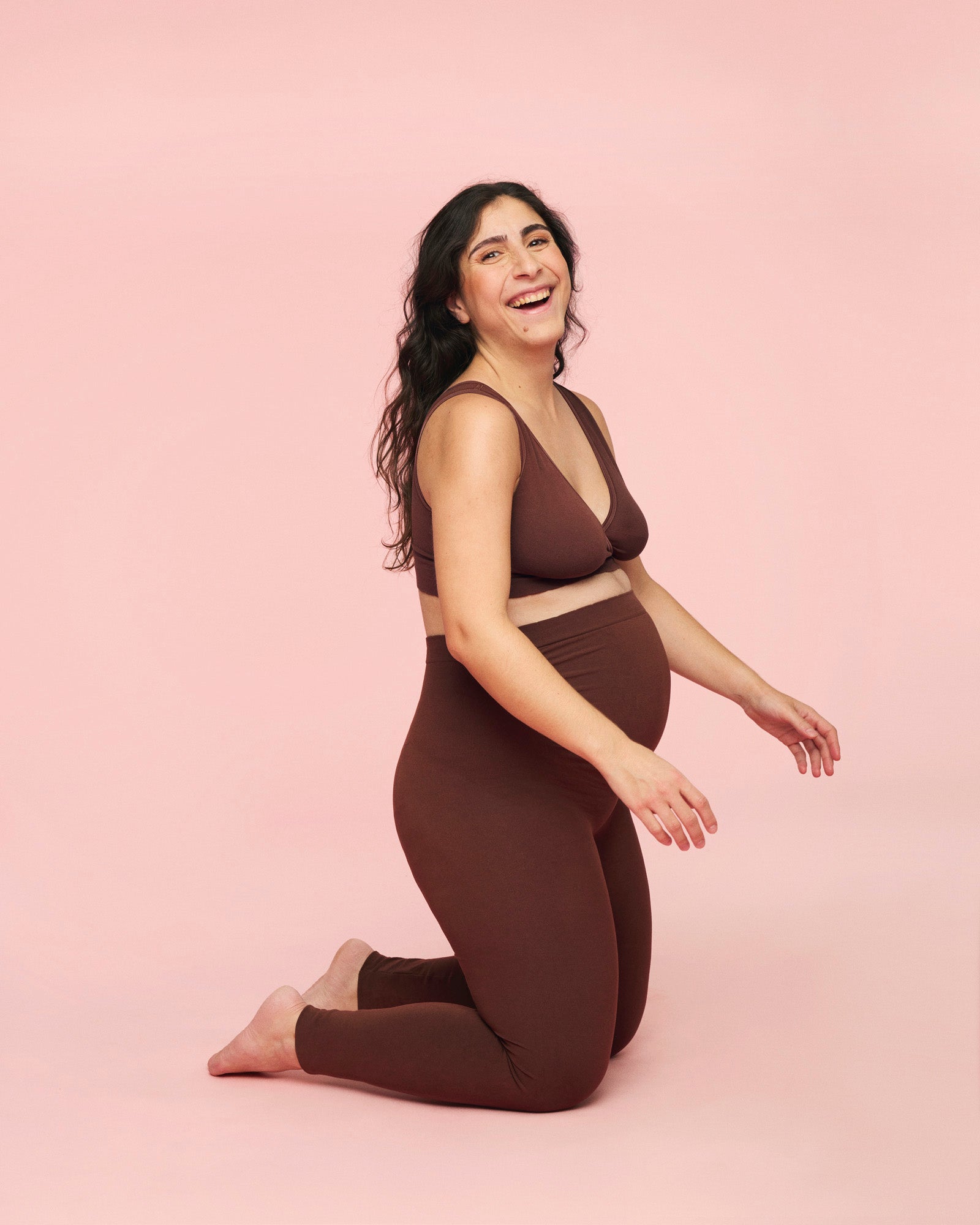 Gravid kvinde sidder på knæ imens hun er iført momkind graviditetssæt med leggings og crossover bh i brun.