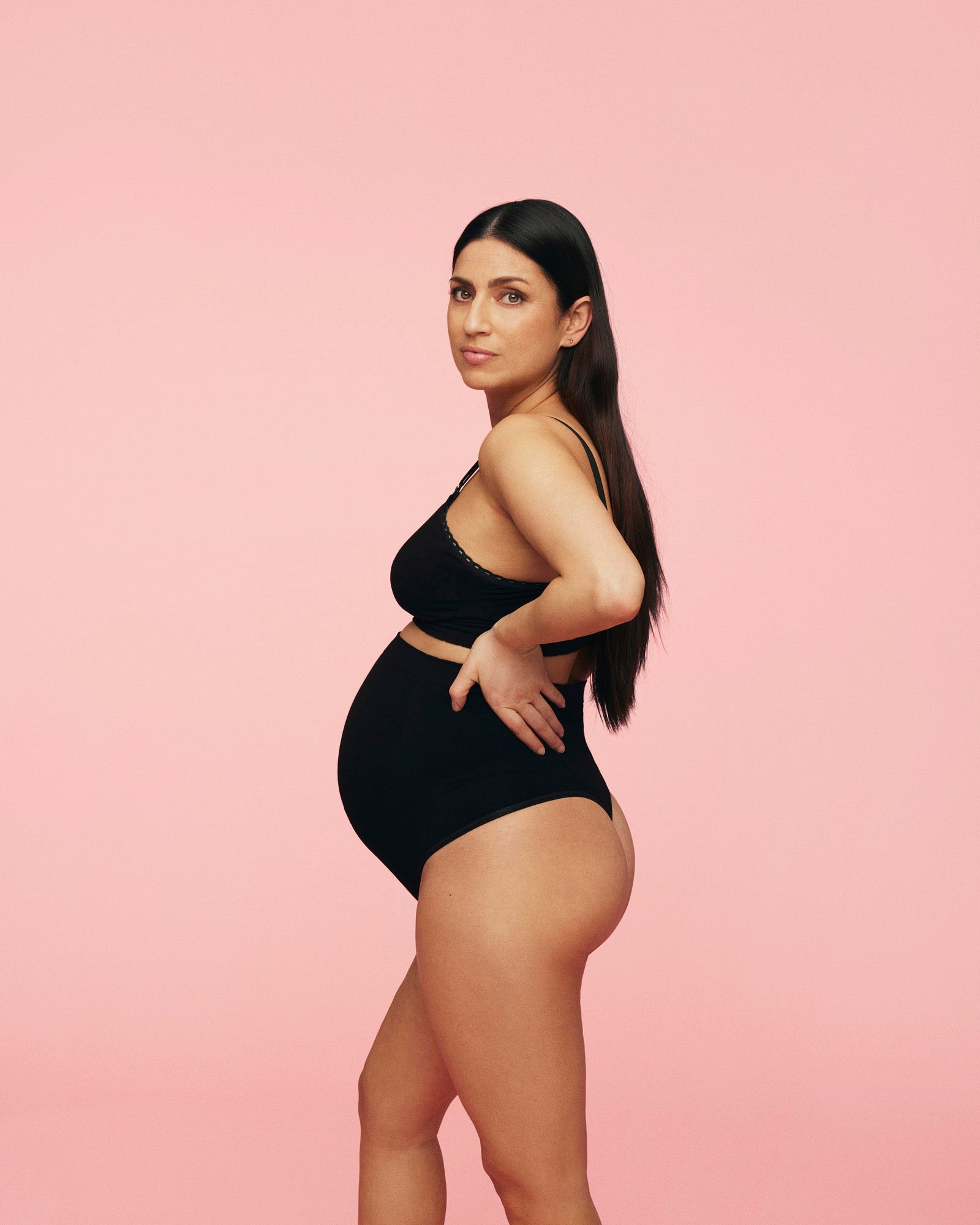 Gravid kvinde iført sort graviditets g-streng med matchende amme bh. Hun står med siden til, med hånden i siden, Foran en rosa baggrund.