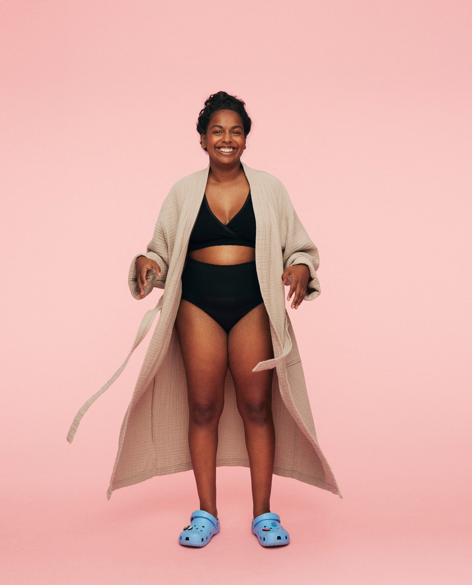 Kvinder smiler imens hun står front mod kameraet iført momkind the kind robe i beige. Den er åben og under har hun momkind trusser og crossover bh på.
