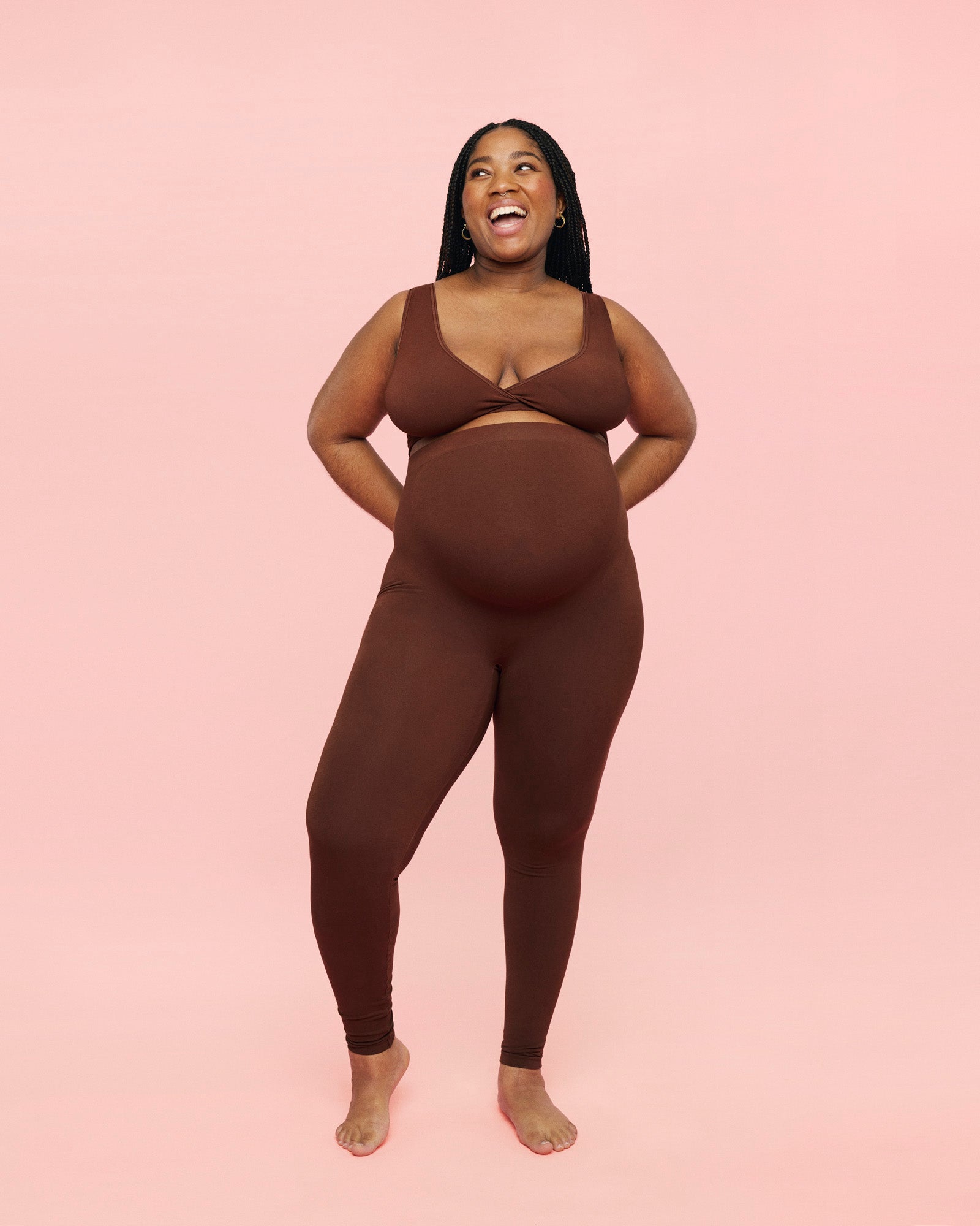 Gravid kvinde iført momkind crossover bra og gravid leggings i farven chocolate. Hun har armene bagved ryggen, så man kan se maven strutte frem, mens hun smiler stort. 