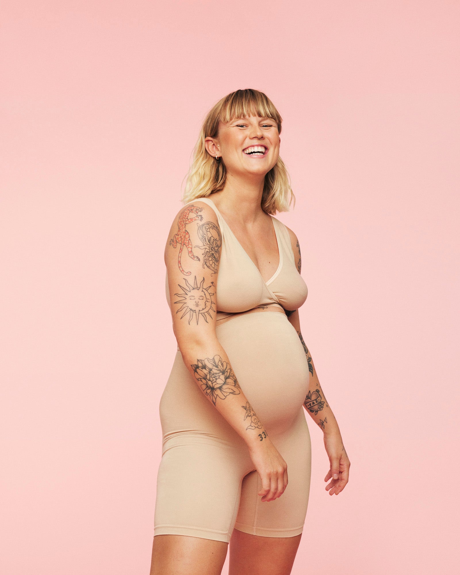 Kvinde iført momkind crossover bra og graviditets shorts i sand. Hun smiler og ser komfortabel ud i tøjet.