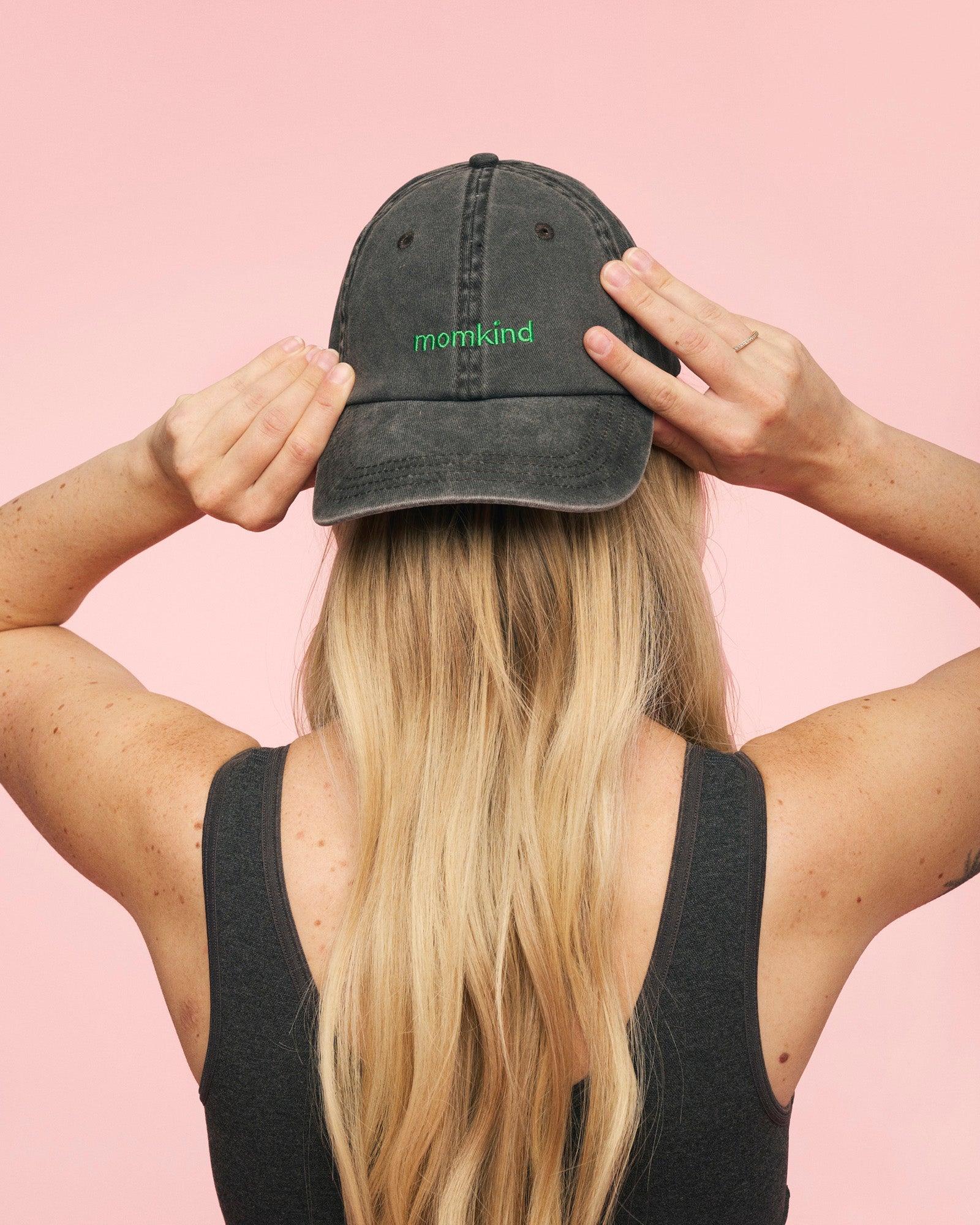 Momkind cap I sort med groent logo. Den sidder omvendt på hovedet af en kvinde som holder begge hænder på.