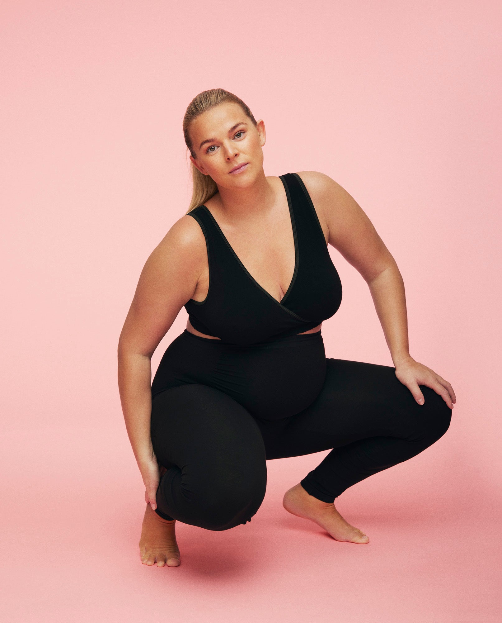 Kvinde sidder på hug mens hun er iført momkind graviditets leggings i sort. Hun er også iført crossover bra i sort. Måden hun sidder på viser at der er stræk i gravid leggings.