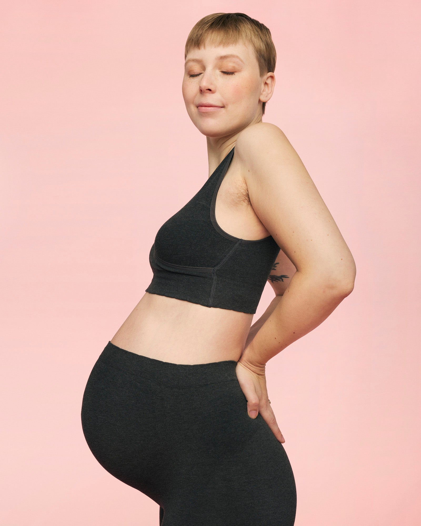 gravid kvinde står med lukkede øjne og hænderne i lænden iført støttende gravid leggings i mørkegrå fra momkind og crossover bh til gravide i grå fra momkind